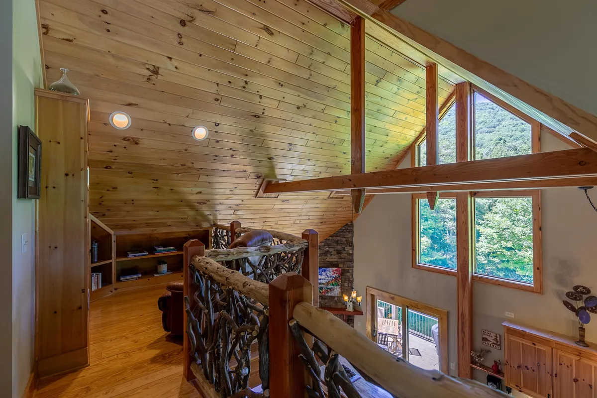 The upstairs reading nook is defined by a wooden railing, shelves filled with books, large windows, and exposed wooden beams. It offers a warm, inviting spot overlooking the living space below.