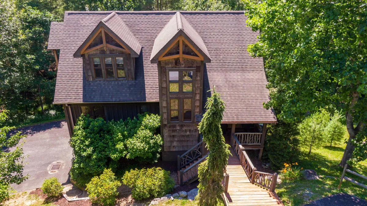 The image showcases the charming exterior of a single-family home surrounded by lush greenery, featuring a welcoming wooden entrance and large front windows. The front porch invites guests to relax and take in the tranquil setting, with hints of sunset hues casting a warm, inviting glow over the property. The scene hints at both the serenity of nature and the comfort of a well-appointed mountain retreat.