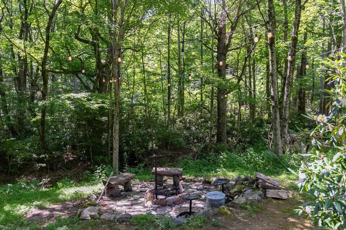 This exterior image showcases a cozy stone fire pit area surrounded by wooden benches, nestled among tall trees. String lights hang above, creating a warm and inviting ambiance. The setting is perfect for gathering with friends or family, telling stories, and enjoying the nighttime outdoors under the stars.