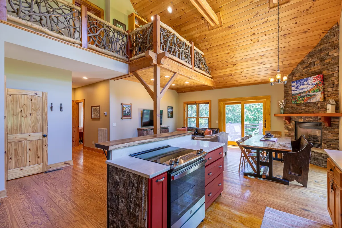 A cozy interior view featuring the kitchen island as a focal point, framed by elegant wooden beams and a striking stone fireplace. Large windows flood the space with natural light, highlighting the open-plan flow between the kitchen and living areas.