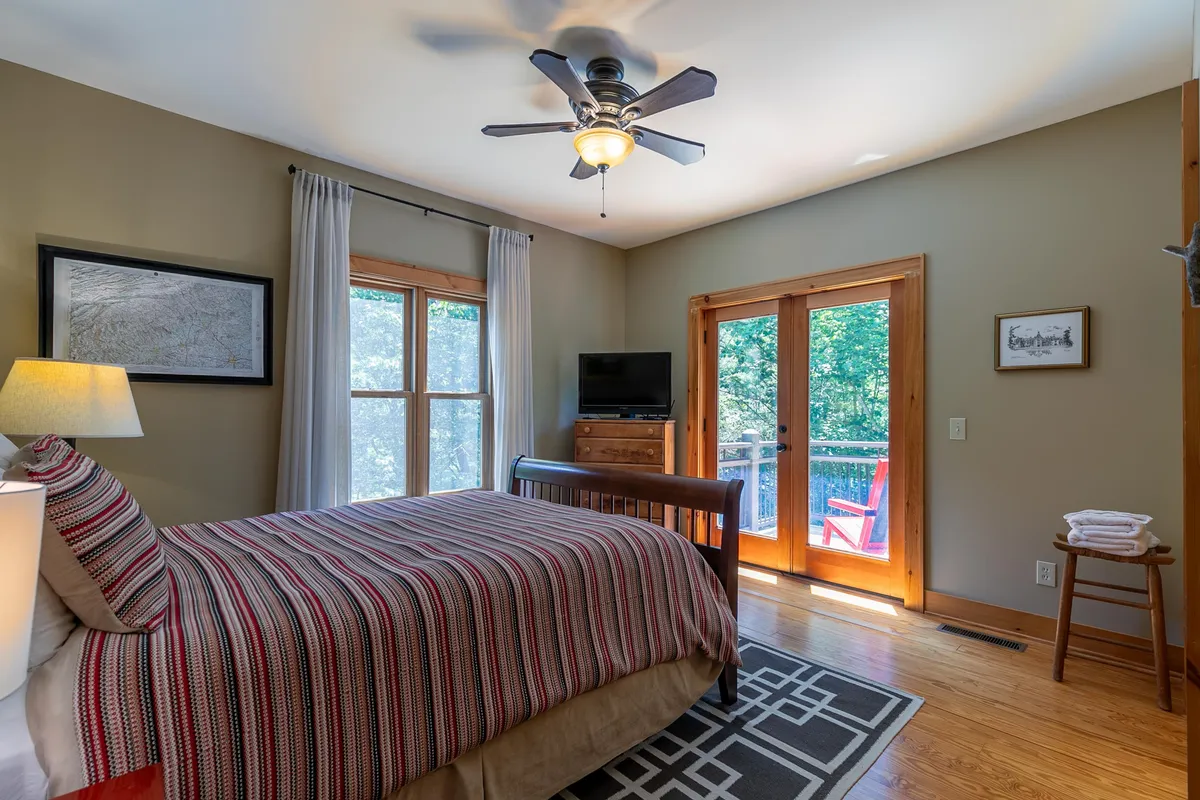 This interior image shows a cozy bedroom with striped bedding, large windows filling the room with natural light, and double doors leading directly to a deck. The wooden furniture adds warmth, while the visible deck just outside the doors hints at easy indoor-outdoor living and opportunities to soak in the surrounding mountain air.