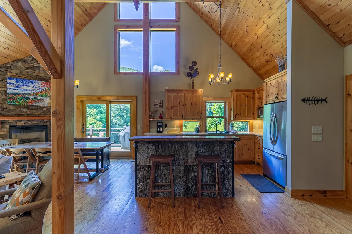 An expansive view of the open-plan kitchen and living area, featuring a spacious kitchen island, wooden beams, and a grand stone fireplace. Floor-to-ceiling windows illuminate the room, creating a bright and welcoming environment ideal for gatherings.