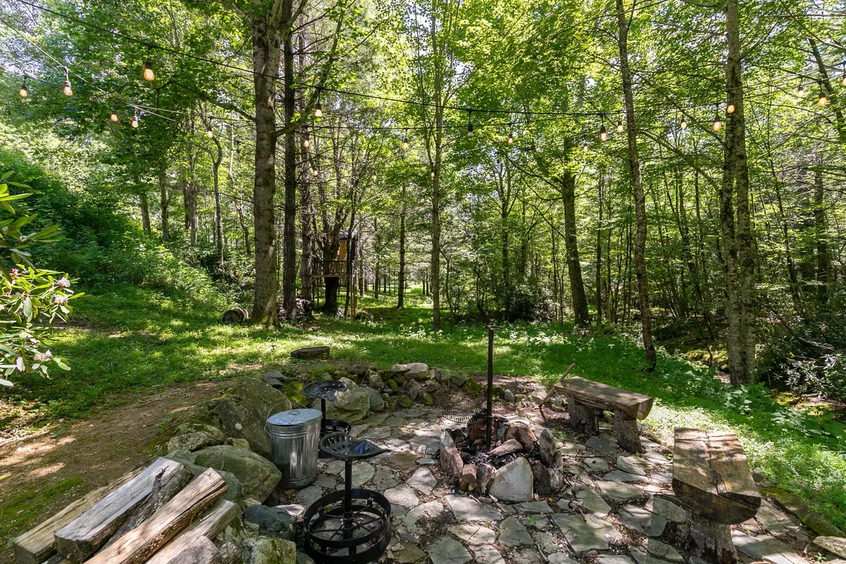 This image captures a cozy backyard setting featuring a stone fire pit surrounded by wooden benches and string lights, with a charming treehouse visible in the background. The setting is embraced by lush greenery, creating a magical atmosphere for evening gatherings and family fun.