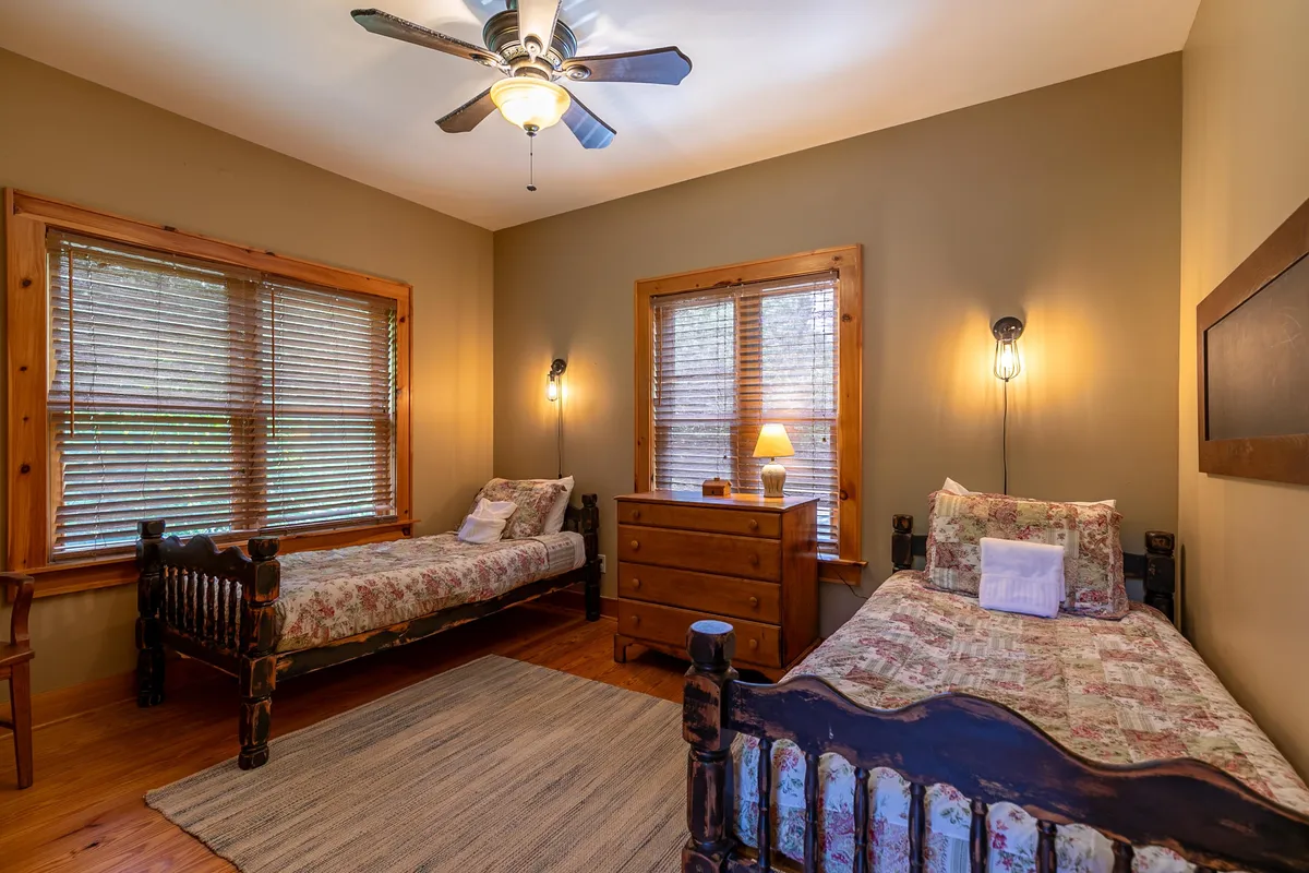 The room shows two wooden beds with floral bedding, a chest of drawers, and large windows with blinds letting in natural light. The space feels warm, inviting, and ideal for children or guests.