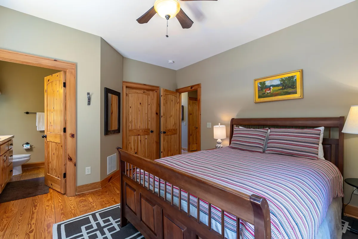 This image features a bed with striped bedding, wooden furniture, a framed painting, and a ceiling fan. The room radiates a cozy, restful vibe with sunlight filtering softly through the windows.