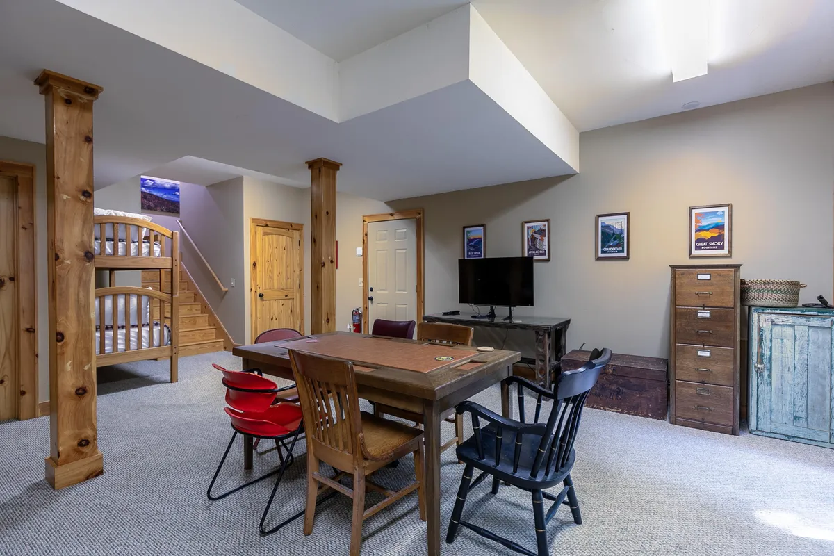 A welcoming game room featuring two bunk beds positioned along the wall, a wooden dining/game table ready for board games or card play, a television unit for movie nights, and ample wooden cabinetry. The room exudes a casual, family-friendly atmosphere ideal for both sleepovers and playful gatherings.